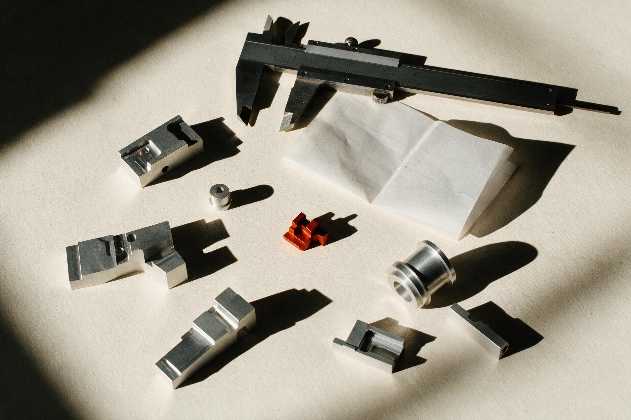 An overhead view of machined aluminium components, a steel caliper and a single orange anodised part arranged on a warm off-white workbench.